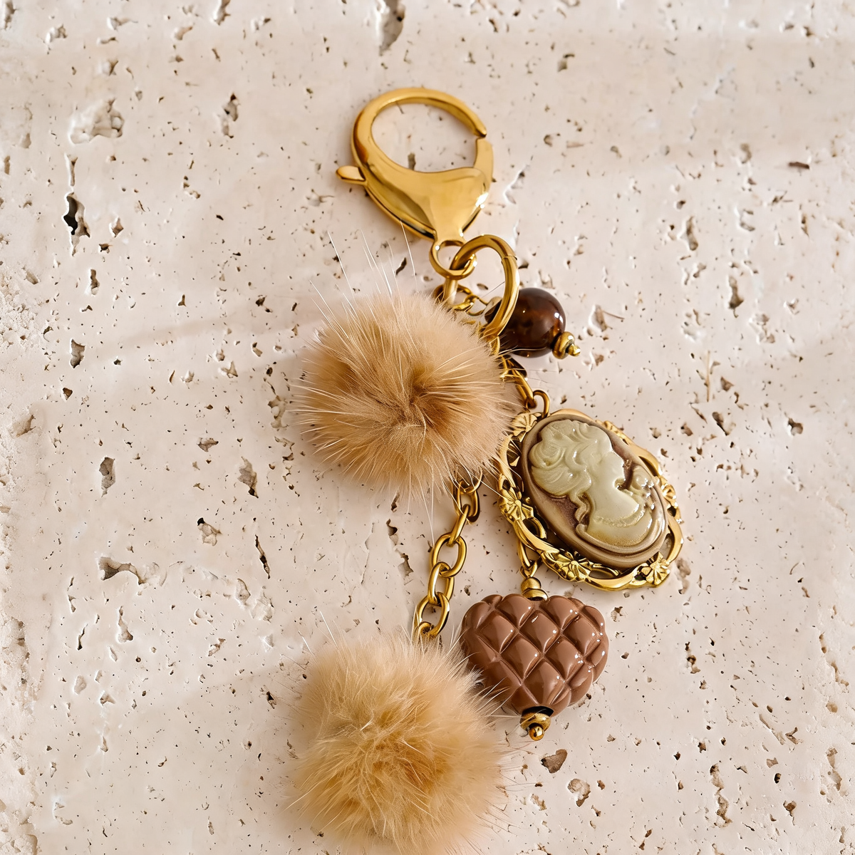 Posy Heart Portrait Keychain in Camel - gold-plated charm keychain with resin cameo, quilted heart and faux fur poms - styled on travertine